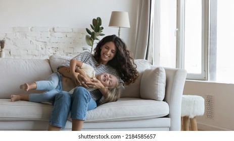 Cheerful happy mom cuddling cute little daughter kid on sofa, tickling, caressing laughing screaming child with love. Mother enjoying activity, leisure with girl playing funny game at home. Banner