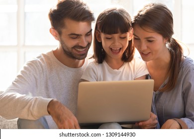 Cheerful Happy Mom Dad And Little Kid Daughter Using Laptop Together At Home, Loving Young Family With Child Girl Laughing Looking At Computer Screen, Enjoying Shopping Or Watching Cartoons Online