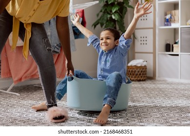 Cheerful Happy Mom Is Playing With Baby In Laundry Room. Little Boy Sits In Clothes Bowl Raises Hands Laughing. Mother Pulls Son For Fun.