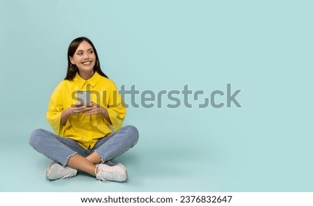 Cheerful happy pretty young woman wearing casual outfit sitting on floor, using cell phone isolated on blue studio background, looking at copy space, checking nice online offer, newest mobile app