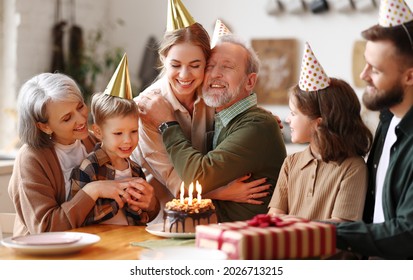 Cheerful happy senior man hugging with his young daughter and feeling thankful while celebrating his birthday with big loving family, sitting together in party hats at table with cake and gift box - Powered by Shutterstock