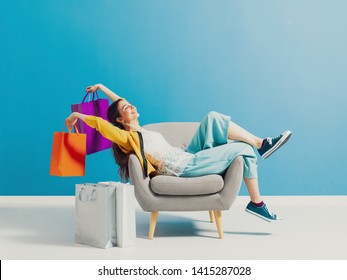 Cheerful happy shopaholic woman with lots of shopping bags, she is sitting on an armchair and celebrating with arms raised