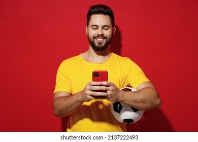 Cheerful happy smiling fun young bearded man football fan in yellow t-shirt cheer up support favorite team hold soccer ball use mobile cell phone isolated on plain dark red background studio portrait