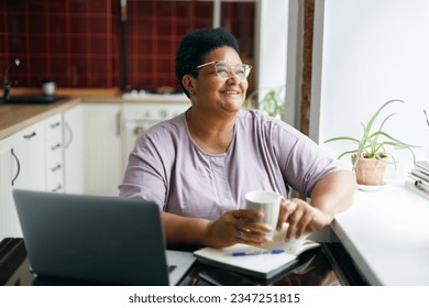 Cheerful happy smiling overweight mature black woman in spectacles sitting at kitchen table with white coffee cup in hands looking through window, enjoying hot drink, after working online on laptop - Powered by Shutterstock