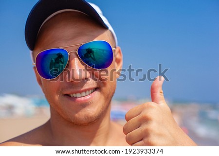 A cheerful and happy tanned man in sunglasses on the beach shows a thumbs up. The reflection in the sunglasses.