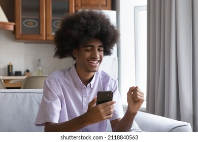 Cheerful Happy Teen African Guy With Curly Fuzzy Hair Excited With Good News In Text Message, Reading Notice On Smartphone Screen, Making Winner Success Yes Hand Gesture On Home Sofa