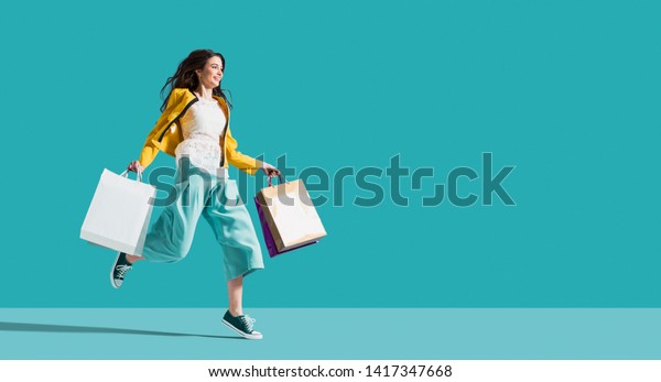 Cheerful happy woman enjoying shopping: she is\
carrying shopping bags and running to get the latest offers at the\
shopping center