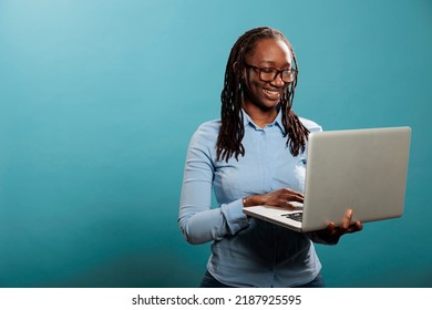 Cheerful Happy Woman Having Laptop And Smiling Heartily While Browsing Webpages On Internet. Joyful Pretty Lady With Handheld Modern Computer Standing On Blue Background While Surfing Browser.