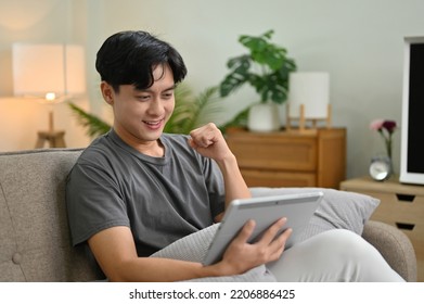 Cheerful and happy young Asian man in casual clothes enjoying his weekend at home, watching a football match on his tablet while relaxing in his living room.