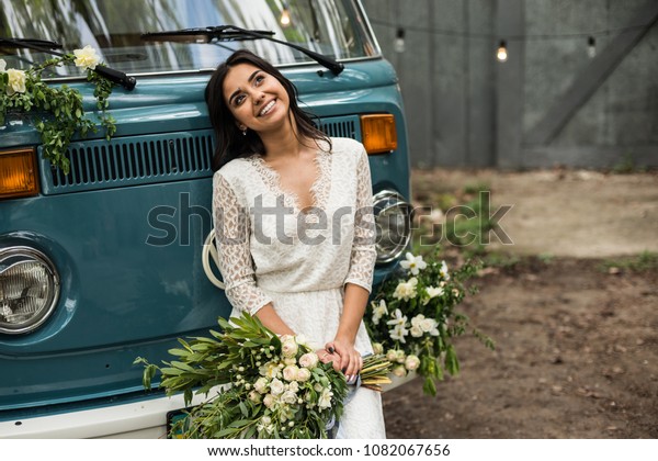 Cheerful happy young bride sit on the bumper\
retro-minibus.\
Close-up.