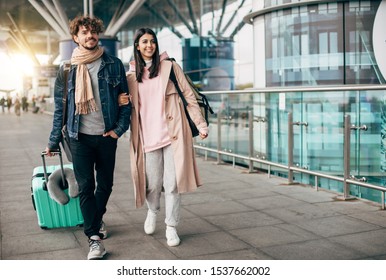 Cheerful happy young couple walk together outside of airport. Guy carry suitcase. She hold his hand. Travel together. Trip or vacation. Sunlight outside. Walking together