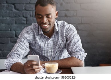 Cheerful and happy young dark-skinned businessman messaging online, checking newsfeed on social networks, using free wireless internet connection during coffee break in modern restaurant interior