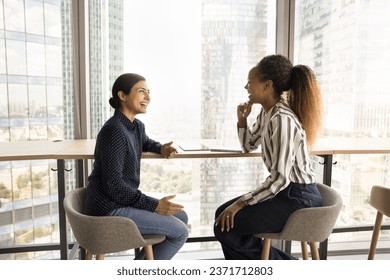 Cheerful happy young diverse office friends girls chatting on work break, discussing job success, talking at large window, laughing, enjoying friendship, networking, professional communication