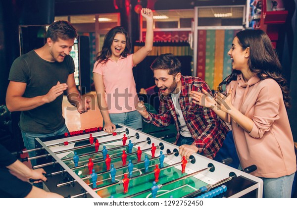 Cheerful happy young team stand at soccer table in playing room and cheering. Bearded guy play with another human. Frieds surround him.