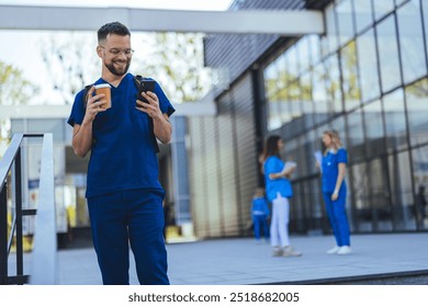 A cheerful healthcare worker in blue scrubs checks his phone while holding coffee outside a modern building. Colleagues are visible in the background, dressed similarly and engaged in conversation. - Powered by Shutterstock