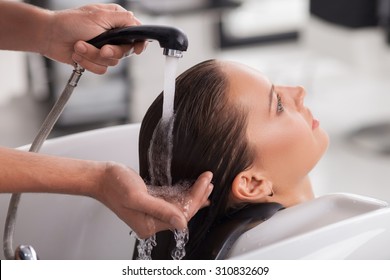 Cheerful healthy girl is getting a hairwash in hair salon. She is sitting and leaning her head on the sink. The lady is relaxing and looking forward with happiness. The beautician is holding a faucet  - Powered by Shutterstock