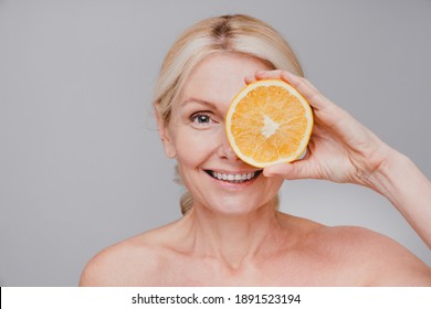 Cheerful healthy mature woman holding orange cut close to her face isolated over grey background