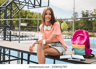 Cheerful high school girl teenager pupil college student in casual clothes with bag sitting in sport yard basketball court holding smart mobile phone doing homework looking at camera - Powered by Shutterstock