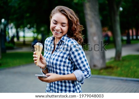 Cheerful hipster girl with modern smartphone technology and sorbet ice cream smiling during weekend summertime spending in park, joyful female holding mobile phone and gelato dessert in hands