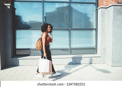 Cheerful Hipster Girl With Shopping Bags In Hands Calling For Boast Of Good Purchases During Black Friday Using Roaming Internet Connection On Modern Smartphone, Full Length With Copy Space Area