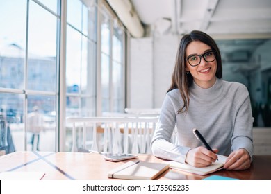 Cheerful hipster girl working as journalist enjoying leisure at cafeteria and looking at camera during break, successful happy writer creating new novel ideas while sitting at coworking space - Powered by Shutterstock