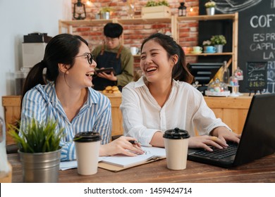 Cheerful Hipster Girls Coworkers In Shirt Laughing While Having Positive Conversation. Two Freelancer Colleagues Using Laptop Computer At Cafe Table. Blurred View Male Waiter In Counter Making Coffee