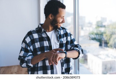 Cheerful Hipster Guy In Casual Clothes Checking Time On Wearable Smartwatch Using 4g Connection Near Window, Happy Male Generation Z Using Digital Wrist Clock While Waiting Colleague In Office