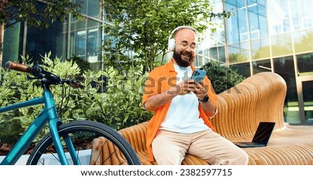 Cheerful hipster man in glasses and headphones usesing smartphone, writes on social networks and sends an online message chating frends, sitting on bench near his bicycle on background of city street.