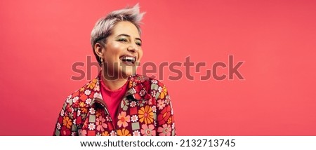 Cheerful hipster woman looking away with a smile on her face in a studio. Happy young woman standing alone against a red background. Fashionable young woman with dyed hair feeling vibrant.