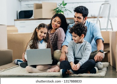 Cheerful Hispanic Family Looking At Laptop While Sitting On Carpet In New Home