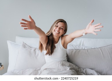 Cheerful hispanic young woman in white t-shirt sits on bed spreads hands welcoming to hug, toothy smiles looks at camera. Happy female at morning feels love tende wants  to embrace. Happy people.