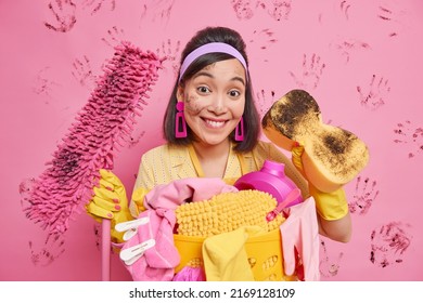 Cheerful Housemaid Holds Dirty Sponge And Mop Does Domestic Chores Cleans House With Lots Of Tools Wears Headband Rubber Gloves Poses Against Pink Background With Handprints. Housework Concept