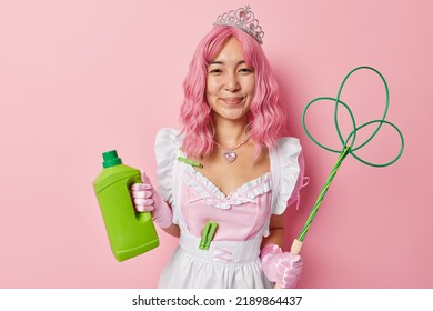 Cheerful Housemaid Wears Crown And White Dress Ready For Cleaning And Washing Holds Green Bottle Of Detergent And Carpetbeater Does Housework Isolated Over Pink Background. Daily Chores And Cleanup