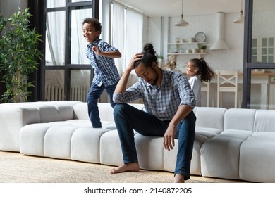 Cheerful Hyperactive Black Sibling Kids Running Around Tired Dad, Jumping On Sofa, Playing Active Games, Screaming, Laughing. Young Father Exhausted With Watching Children, Feeling Headache, Stress