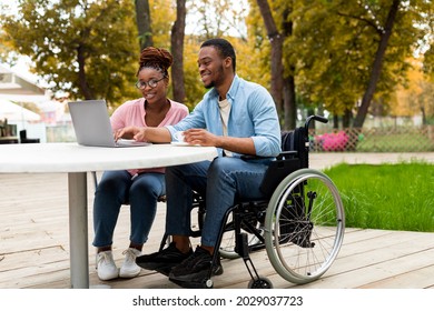 Cheerful Impaired Black Man With Female Colleague Using Laptop Together In Outdoor Cafe At Autumn Park, Working On Online Business Project, Attending Meeting Remotely