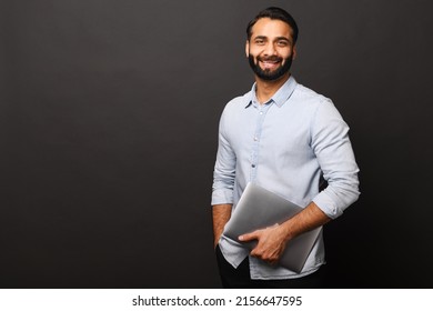 Cheerful Indian Bearded Man Holding Laptop Standing Isolated On Black, Looking At Camera With Happy Smile. Male Employee Or Freelancer Carrying Laptop Computer. Copy Space