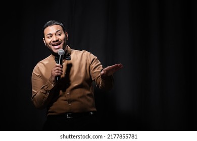 Cheerful Indian Comedian In Shirt And Bow Tie Holding Microphone During Monologue On Black