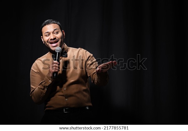 cheerful indian comedian in shirt and\
bow tie holding microphone during monologue on\
black