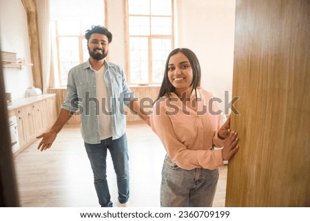 Cheerful indian couple inviting guests to enter home, happy people standing in doorway of modern flat, looking out, waiting for visitor to come in, receiving friend