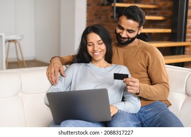Cheerful Indian couple watching at laptop screen and checking credit card number to make online transaction. Smiling spouses having shopping online. Transferring money, online banking concept
