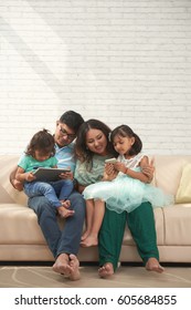 Cheerful Indian Family Sitting On Sofa With Smartphone And Digital Tablet