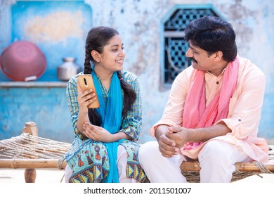 Cheerful Indian Father And Young Daughter Using Smartphone Looking At Each Other While Sitting On Traditional Wooden Bed Outside Their House, Happy Rural Family, Technology Concept.  