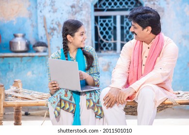 Cheerful Indian Father And Young Daughter Using Laptop Looking At Each Other While Sitting On Traditional Wooden Bed Outside Their House, Happy Rural Family, Web Surfing, Technology Concept.        