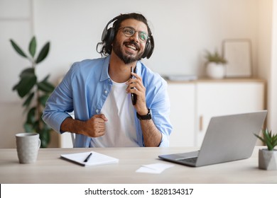 Cheerful indian freelancer guy singing and listening music with headphones while working with laptop at home office, using smartphone as microphone, enjoying his favorite song, copy space - Powered by Shutterstock