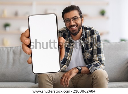 Cheerful Indian Guy Showing Smartphone With Big Blank White Screen At Camera, Happy Young Eastern Man Recommending New Mobile App Or Website While Sitting On Couch At Home, Creative Collage, Mockup