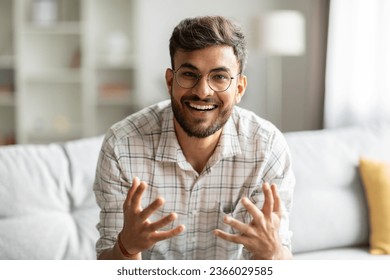 Un chico indio alegre hablando mirando la cámara y haciendo gestos, comunicándose en línea sentado en el sofá en casa. Vista de cámara web del hombre charlando de forma remota a través de videollamadas