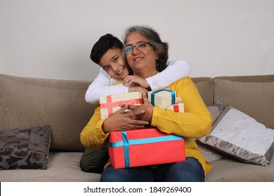 Cheerful Indian Kid And His Grand Mother Sitting On Sofa With Gifts.