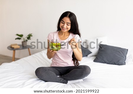 Cheerful Indian lady sitting on bed at home with bowl of fresh vegetable salad, full length. Pretty Asian woman enjoying vegetarian meal. Clean living, healty eating, vegan diet concept