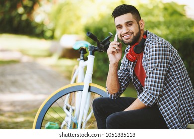 Cheerful Indian Man Looking At Camera With Toothy Smile While Talking To Friend On Smartphone And Sitting On Park Bench, Modern Bicycle On Background