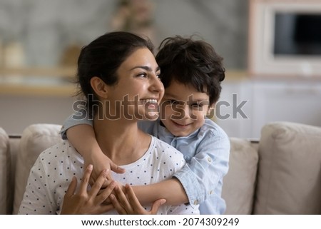 Cheerful Indian preschool kid hugging happy mom with love, gratitude, tenderness. Young mother and little son enjoying leisure time together, looking away, smiling. Family, motherhood concept
